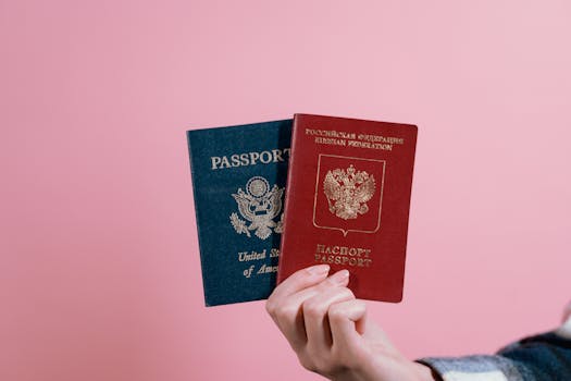 Close-up of a hand holding American and Russian passports on a pink background.